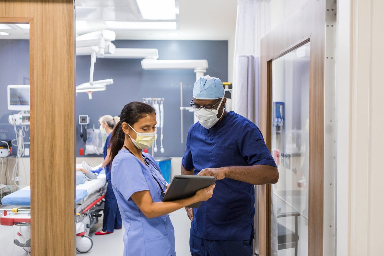 As a mature nurse monitors the patient on gurney, the mature adult female nurse shows the mature adult male doctor the test results on a digital tablet.