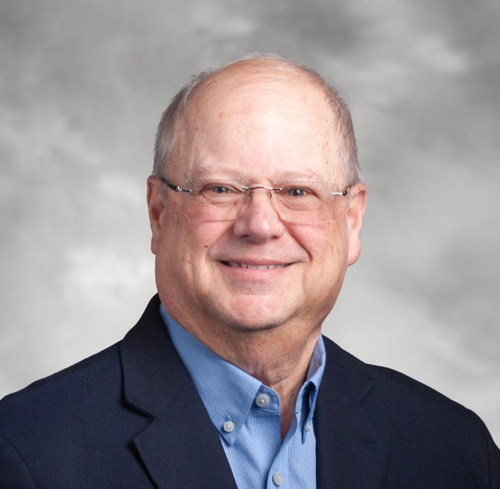 Smiling man in a blue shirt and dark blazer against a neutral background.